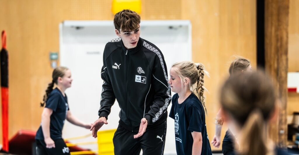 Training-12_Titel Ein Trainer in einem schwarzen Puma-Trainingsanzug spricht während eines Hallentrainings mit einem jungen Mädchen in Sportkleidung, während im Hintergrund andere Kinder aktiv sind.