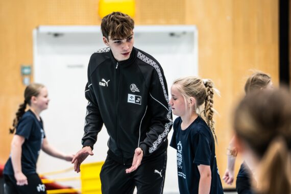 Ein Trainer in einem schwarzen Puma-Trainingsanzug spricht während eines Hallentrainings mit einem jungen Mädchen in Sportkleidung, während im Hintergrund andere Kinder aktiv sind.