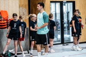 Ein Trainer steht in einer Sporthalle, während sich eine Gruppe junger Mädchen in Sportkleidung auf das Training vorbereitet; einige halten Basketbälle in der Hand.