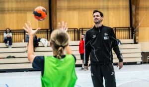 Ein Trainer in einem schwarzen Trainingsanzug beobachtet, wie ein junger Sportler einen orangefarbenen Handball in einer Sporthalle wirft, im Hintergrund sind leere Tribünen zu sehen.