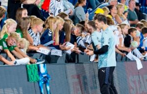 Ein Fußballspieler steht neben der Tribüne und gibt begeisterten Kindern, die Trikots seiner Mannschaft tragen, Autogramme, während im Hintergrund eine Menschenmenge zusieht.