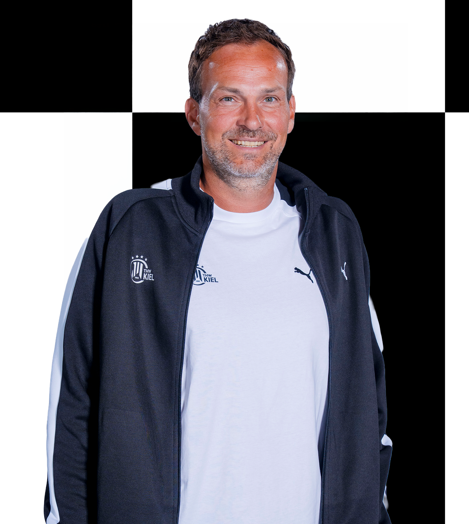 Ein lächelnder Mann, der ein schwarz-weißes Sakko über einem weißen Hemd trägt, steht vor einem schwarz-weiß karierten Hintergrund, der die Geschäftsstelle des THW Kiel symbolisiert. Er steht Ihnen bei allen Anfragen bezüglich Mitarbeiterkontakten gerne zur Verfügung.
