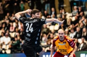 Ein Handballspieler im schwarzen Trikot macht sich bereit, den Ball zu werfen, während ein Verteidiger im rot-gelben Trikot ihn aufmerksam beobachtet. Die Zuschauer im Hintergrund sind verschwommen.