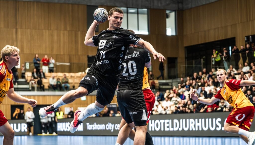 gog_elis Ein Handballspieler in schwarzer Spielkleidung springt zum Wurf ab, während ein Verteidiger versucht, den Wurf zu blocken. Andere Spieler in Rot und Gelb schauen zu, und die Zuschauerränge einer modernen Halle sind voll besetzt.