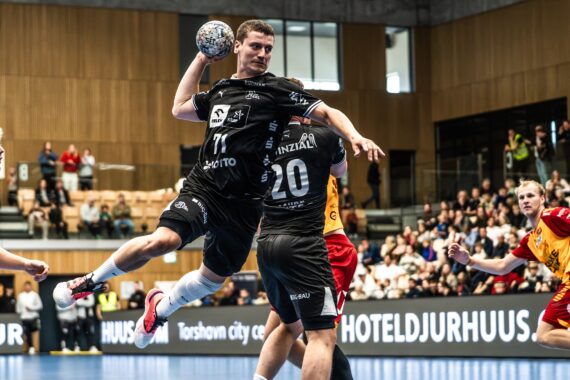 Ein Handballspieler in schwarzer Spielkleidung springt zum Wurf ab, während ein Verteidiger versucht, den Wurf zu blocken. Andere Spieler in Rot und Gelb schauen zu, und die Zuschauerränge einer modernen Halle sind voll besetzt.