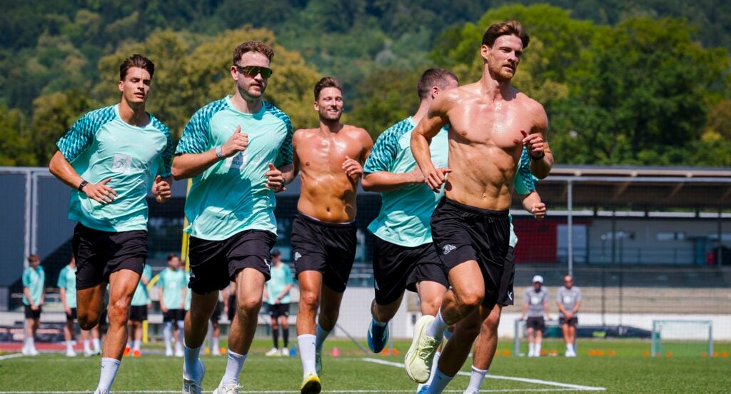Eine Gruppe männlicher Fußballspieler, von denen einige ohne Trikot sind, läuft während des Trainings gemeinsam auf einem Außenplatz, im Hintergrund sind Bäume und ein Stadion zu sehen.
