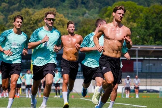 Eine Gruppe männlicher Fußballspieler, von denen einige ohne Trikot sind, läuft während des Trainings gemeinsam auf einem Außenplatz, im Hintergrund sind Bäume und ein Stadion zu sehen.