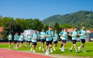 Eine Gruppe männlicher Fußballspieler in einheitlichen hellblauen Trikots joggt gemeinsam auf einer Leichtathletikbahn, im Hintergrund sind grüne Bäume, Hügel und Häuser vor einem strahlend blauen Himmel zu sehen.
