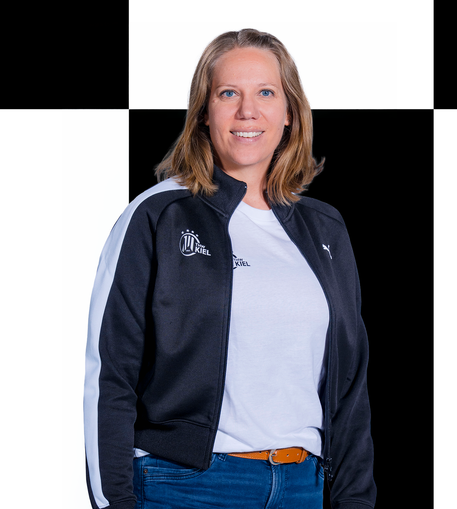 Eine Frau mit schulterlangen blonden Haaren, eine der Mitarbeiterinnen der THW Kiel-Geschäftsstelle, trägt eine schwarz-weiße Sportjacke und ein weißes Hemd, steht vor einem schwarz-weiß karierten Hintergrund und lächelt in die Kamera. Die Kontaktdaten finden Sie im offiziellen Teamverzeichnis.