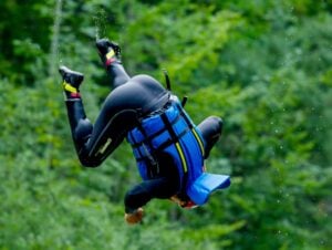 Eine Person, die eine blaue Schwimmweste und einen schwarzen Neoprenanzug trägt, vollführt gerade einen Salto über dem Wasser, während im Hintergrund grünes Laub verschwommen zu sehen ist.