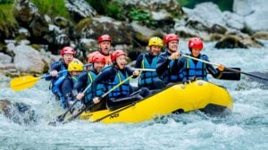 Sieben Personen, die Helme und Schwimmwesten tragen, paddeln mit einem gelben Schlauchboot durch Stromschnellen, umgeben von Felsen und grüner Natur, und wirken dabei begeistert und konzentriert.