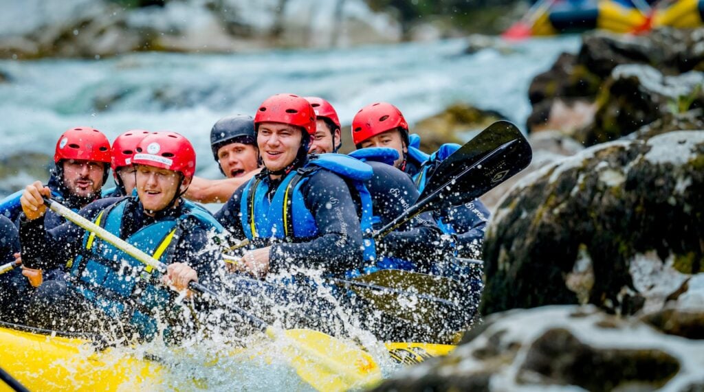 rafting_Titel Eine Gruppe von Menschen mit roten Helmen und Schwimmwesten paddelt in einem gelben Schlauchboot durch spritzende Stromschnellen, umgeben von Felsen und tosendem Wasser.
