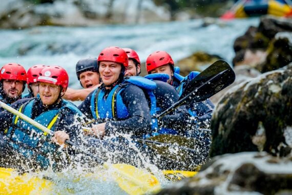 Eine Gruppe von Menschen mit roten Helmen und Schwimmwesten paddelt in einem gelben Schlauchboot durch spritzende Stromschnellen, umgeben von Felsen und tosendem Wasser.