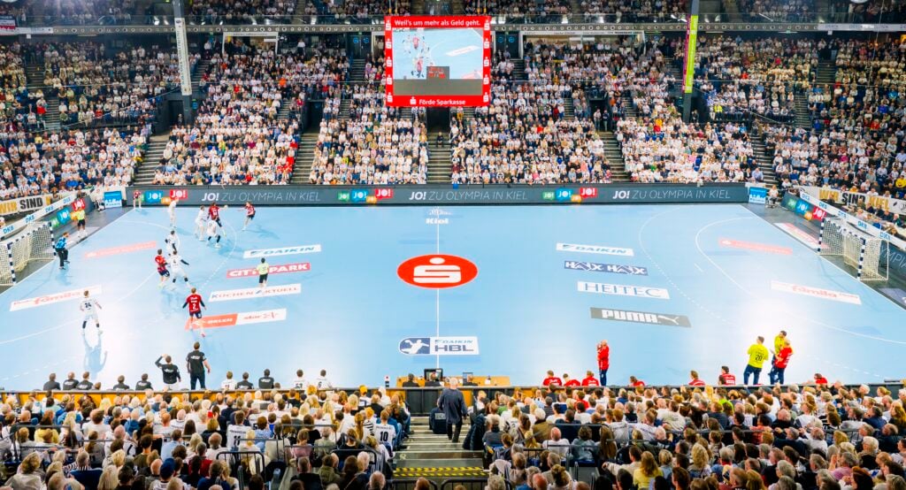 HANDBALL 2025/26: THW Kiel - SG Flensburg-Handewitt Eine große Sporthalle, gefüllt mit Zuschauern, die ein Handballspiel auf einem blauen Spielfeld verfolgen. Spieler in weißen und schwarzen Trikots treten gegeneinander an, über ihnen befindet sich eine Anzeigetafel, und helle Scheinwerfer beleuchten die Szene.