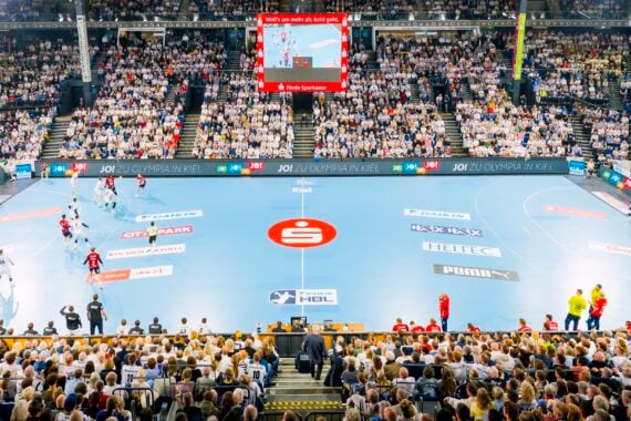 Eine große Sporthalle, gefüllt mit Zuschauern, die ein Handballspiel auf einem blauen Spielfeld verfolgen. Spieler in weißen und schwarzen Trikots treten gegeneinander an, über ihnen befindet sich eine Anzeigetafel, und helle Scheinwerfer beleuchten die Szene.
