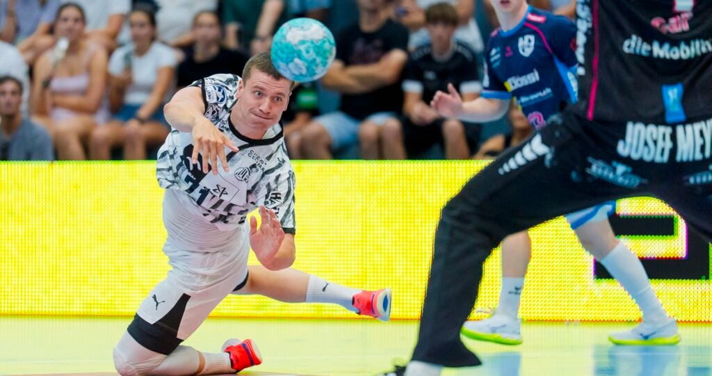 Ein Handballspieler in einem schwarz-weißen Trikot hechtet während eines Spiels in die Luft und wirft den Ball, während Zuschauer zuschauen und im Hintergrund andere Spieler zu sehen sind.