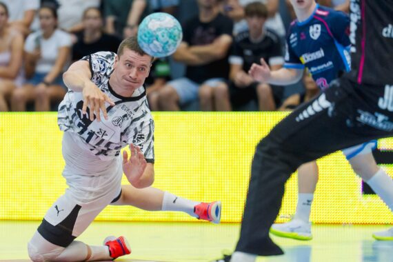 Ein Handballspieler in einem schwarz-weißen Trikot hechtet während eines Spiels in die Luft und wirft den Ball, während Zuschauer zuschauen und im Hintergrund andere Spieler zu sehen sind.