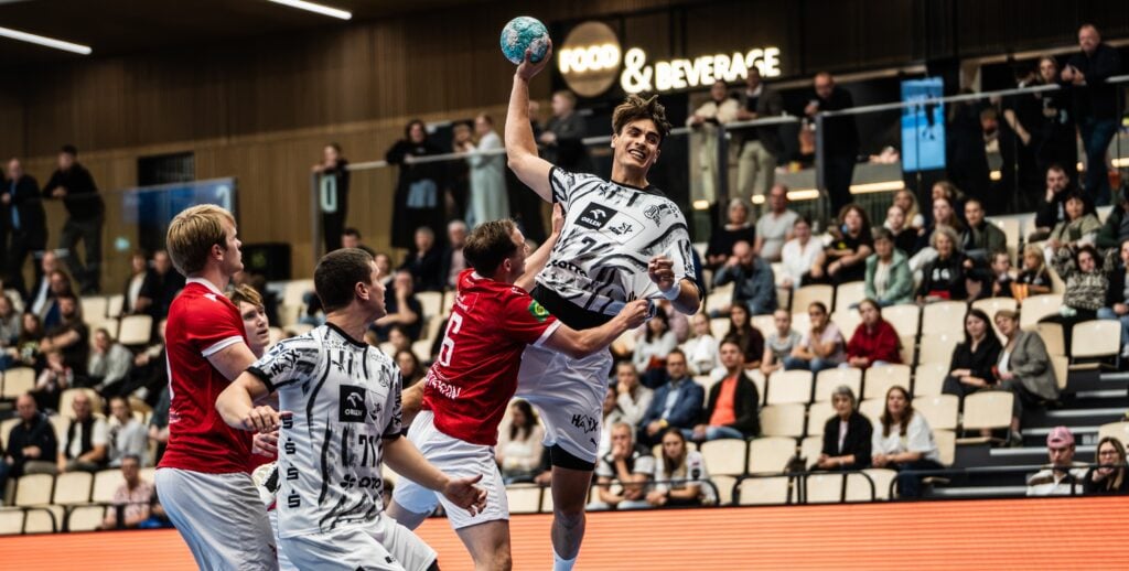 valur1 Ein Handballspiel der Herren zeigt einen Spieler in einem weißen Trikot, der zum Wurf anspringt, während Verteidiger in Rot versuchen, ihn zu blocken; im Hintergrund sind sitzende Zuschauer und ein „Food & Beverage“-Schild zu sehen.