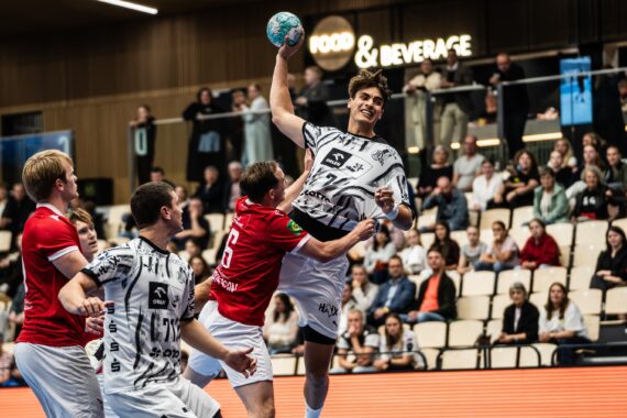 Ein Handballspiel der Herren zeigt einen Spieler in einem weißen Trikot, der zum Wurf anspringt, während Verteidiger in Rot versuchen, ihn zu blocken; im Hintergrund sind sitzende Zuschauer und ein „Food & Beverage“-Schild zu sehen.