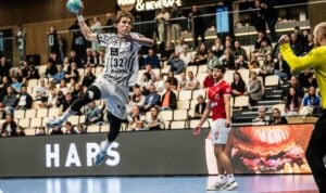 Ein Handballspieler im weißen Trikot springt in der Luft hoch, um einen Wurf abzugeben, und zielt dabei auf das Tor, das von einem Spieler in Gelb verteidigt wird. Zuschauer verfolgen das Geschehen von den Tribünen aus, während ein weiterer Spieler in Rot in der Nähe steht.