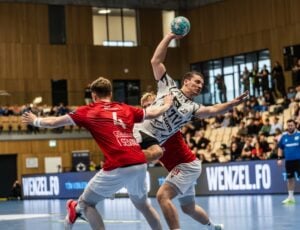Ein Handballspieler in einem weißen Trikot springt hoch, um den Ball zu werfen, während er bei einem Hallenspiel von Gegnern in roten Trikots geblockt wird; Zuschauer verfolgen das Geschehen von den Tribünen aus.