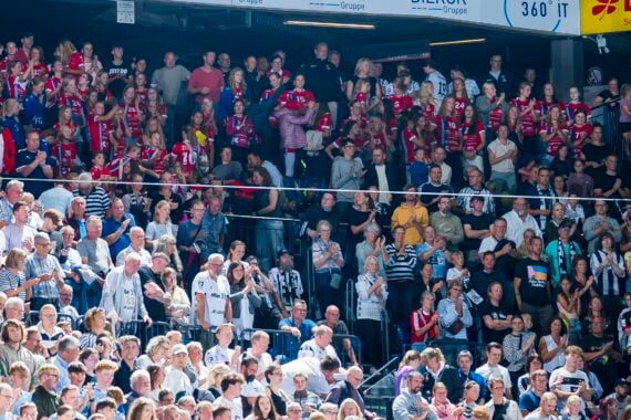Eine große Menge von Fußballfans, von denen einige rote, andere weiße oder schwarze Trikots tragen, jubelt und verfolgt ein Spiel in einem Stadion, über dem Transparente zu sehen sind.
