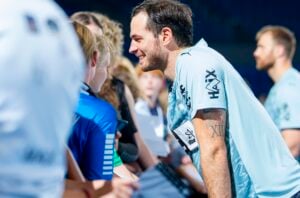 Ein lächelnder Sportler in einem hellblauen Trikot unterhält sich bei einer Hallenveranstaltung mit Fans, gibt Autogramme und geht auf das Publikum ein.