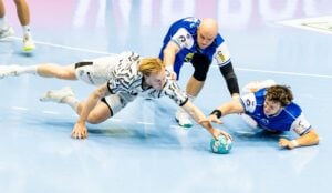 Zwei Handballspieler, einer in Weiß und einer in Blau, stürzen sich auf einem strahlend blauen Spielfeld vor und greifen beide nach einem türkisfarbenen Ball, während sich ein weiterer, blau gekleideter Spieler in den Kampf einmischt.