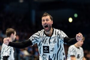 Ein Handballspieler in einem schwarz-weißen Trikot jubelt während eines Spiels mit geballten Fäusten, im Hintergrund sind seine Mannschaftskameraden und eine verschwommene Menschenmenge zu sehen.