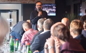 Ein Mann steht vor einem sitzenden Publikum bei einer Veranstaltung in einem Saal, auf den Tischen stehen Getränke, und spricht in ein Mikrofon.