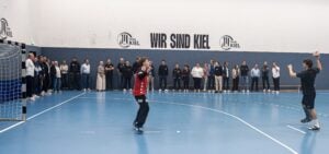 Ein Handballspieler in schwarzer Kleidung macht sich bereit, auf das Tor zu werfen, während der Torwart wartet. Hinter ihm stehen Zuschauer an der Wand, im Hintergrund sind die Aufschrift „WIR SIND KIEL“ und das THW-Kiel-Logo zu sehen.