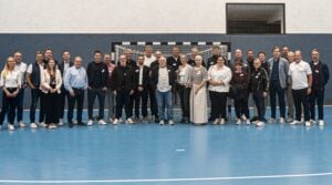 Eine Gruppe von etwa 30 Erwachsenen steht in einer einzigen Reihe auf dem blauen Boden einer Sporthalle und posiert lächelnd vor einem Handballtor mit Netz. Die meisten sind leger gekleidet.