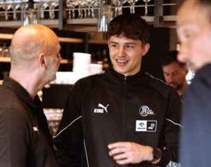 Zwei Männer in schwarzen Sportjacken unterhalten sich lächelnd in einem Restaurant oder Café, über ihnen hängen Weingläser, im Hintergrund sind Regale zu sehen. Ein Mann blickt in die Kamera, der andere steht mit dem Rücken zur Kamera.