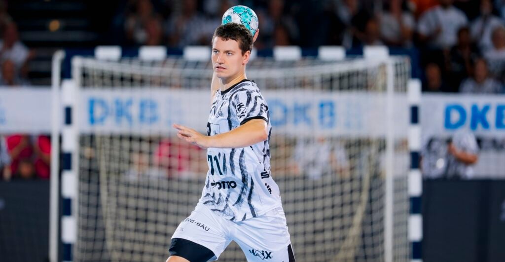 Ein männlicher Handballspieler in einem schwarz-weißen Trikot springt in die Höhe und macht sich bereit, einen blaugrünen Ball zu werfen; im Hintergrund sind ein Tor und verschwommene Zuschauer zu sehen.