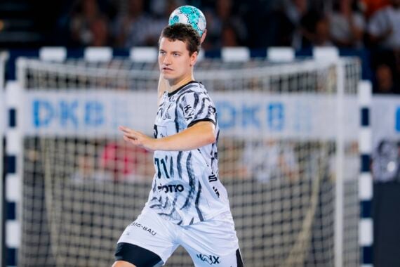 Ein männlicher Handballspieler in einem schwarz-weißen Trikot springt in die Höhe und macht sich bereit, einen blaugrünen Ball zu werfen; im Hintergrund sind ein Tor und verschwommene Zuschauer zu sehen.