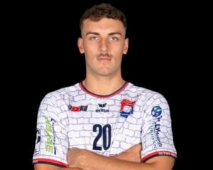 Ein männlicher Sportler mit kurzen Haaren und einem Schnurrbart steht mit verschränkten Armen da; er trägt ein weißes Sporttrikot mit der Nummer 20 und verschiedenen Logos vor schwarzem Hintergrund.