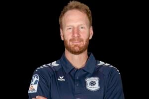Ein Mann mit kurzen roten Haaren und Bart trägt ein marineblaues Handball-Mannschaftstrikot mit Logos. Er steht mit verschränkten Armen vor einem schwarzen Hintergrund und blickt in die Kamera.
