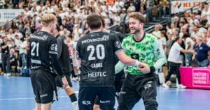 Drei männliche Handballspieler in schwarzen Trikots feiern auf einem Hallenfeld, lächeln und geben sich die Hand. Im Hintergrund beobachtet eine Gruppe von Zuschauern in weißen Hemden das Geschehen von den Tribünen aus.