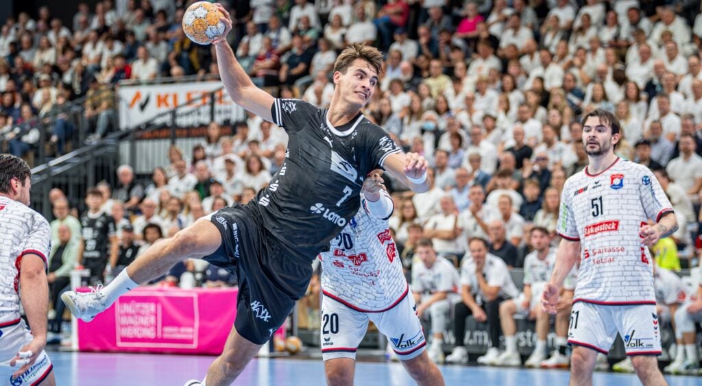 Ein Handballspieler in schwarzer Spielkleidung springt zum Wurf hoch, während Verteidiger in weißer Spielkleidung versuchen, ihn zu blocken. Das Publikum verfolgt das Geschehen in einer vollbesetzten Halle.