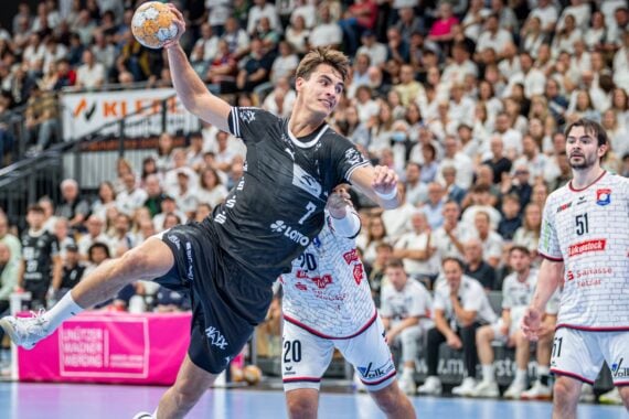 Ein Handballspieler in schwarzer Spielkleidung springt zum Wurf hoch, während Verteidiger in weißer Spielkleidung versuchen, ihn zu blocken. Das Publikum verfolgt das Geschehen in einer vollbesetzten Halle.