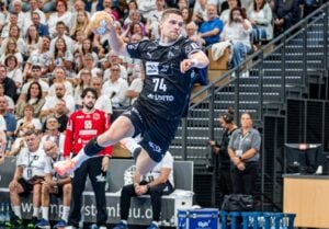 Ein Handballspieler in schwarzer Spielkleidung springt in die Luft, um den Ball zu werfen, während das zahlreich erschienene Publikum und der gegnerische Torwart in Rot das Geschehen während des Spiels gespannt verfolgen.