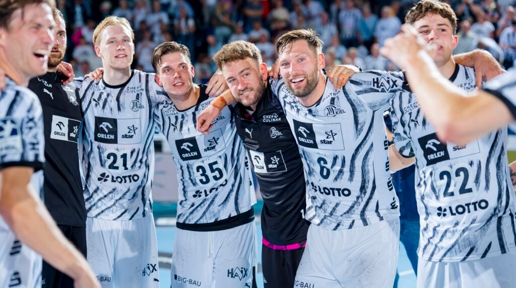 HANDBALL 2026/27: THW Kiel - TBV Lemgo Lippe Eine Gruppe männlicher Sportler in einheitlichen weiß-schwarzen Trikots lächelt und posiert Arm in Arm auf einem Hallenplatz, während sie gemeinsam feiern – im Hintergrund ist eine Menschenmenge zu sehen.