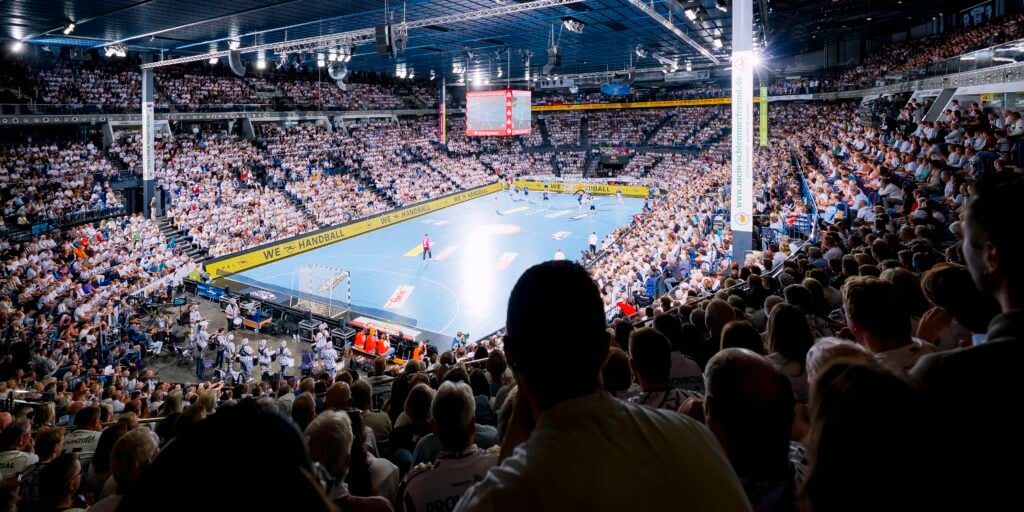Eine vollbesetzte Sporthalle, in der Fans ein professionelles Handballspiel auf einem blauen Spielfeld verfolgen, über ihnen leuchten helle Scheinwerfer, und im Hintergrund ist eine große Anzeigetafel zu sehen.