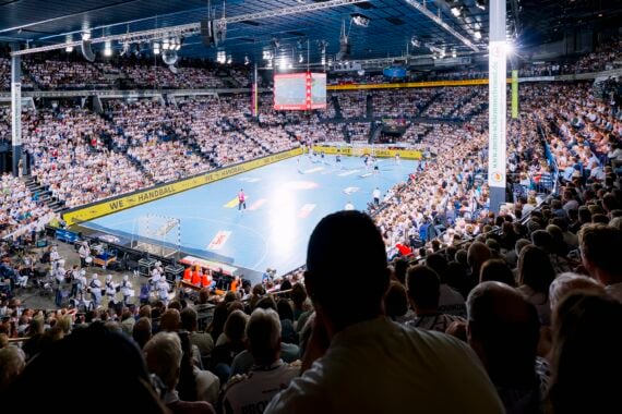 Eine vollbesetzte Sporthalle, in der Fans ein professionelles Handballspiel auf einem blauen Spielfeld verfolgen, über ihnen leuchten helle Scheinwerfer, und im Hintergrund ist eine große Anzeigetafel zu sehen.
