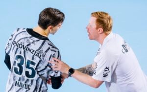 Ein Trainer in einem weißen Hemd spricht intensiv mit einem Spieler, der ein Trikot mit der Rückennummer 35 und dem Namen „Makuc“ trägt. Der Trainer hält den Arm des Spielers fest, und beide befinden sich auf einem hellblauen Sportplatz, wo sie sich auf den Vorbericht zum Auswärtsspiel des THW Kiel beim deutschen Meister SC Magdeburg am Mittwoch vorbereiten.