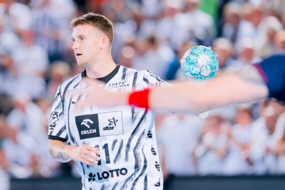 Ein Handballspieler in einem schwarz-weiß gestreiften Trikot hält einen blauen Ball in der Hand, im Vordergrund ist der verschwommene Arm eines anderen Spielers zu sehen, im Hintergrund eine Zuschauermenge.
