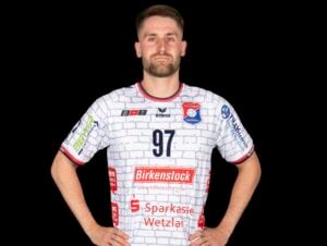 Ein männlicher Sportler, der ein weißes Handballtrikot mit der Nummer 97 und mehreren Sponsorenlogos trägt, steht mit den Händen in die Hüften gestemmt vor einem schwarzen Hintergrund.