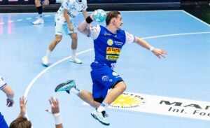 Ein männlicher Handballspieler in einem blauen Trikot springt in die Luft und bereitet sich darauf vor, den Ball zu werfen. Weitere Spieler in weißen Trikots sind zu sehen, und die Szene spielt sich auf einem Handball-Hallenplatz ab.