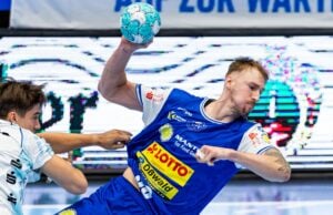 Ein männlicher Handballspieler in einem blauen Trikot springt in die Höhe, um den Ball zu werfen, während ein Gegner in einem weißen Trikot versucht, ihn während eines Spiels zu blocken.