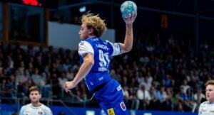Ein männlicher Handballspieler in einem blauen Trikot springt in die Luft, um den Ball zu werfen. Im Hintergrund sind das Spielfeld und die Zuschauer zu sehen.